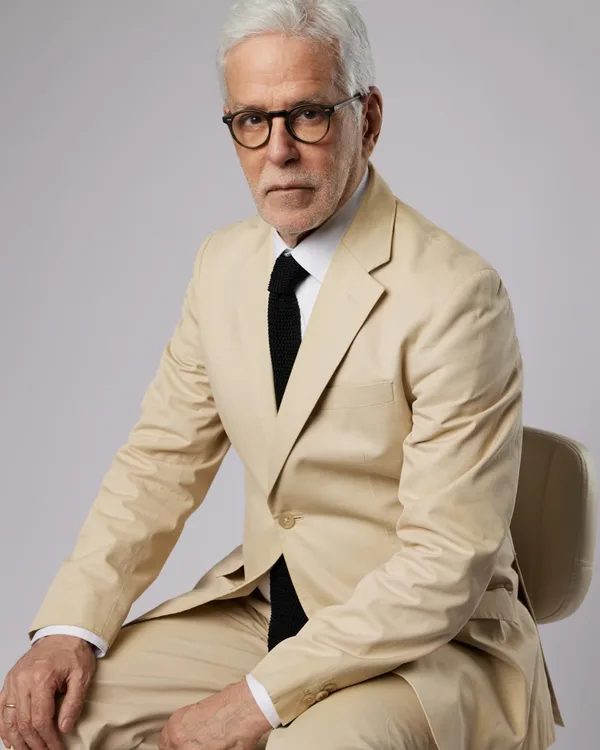 John Amedro seated in a cream suit and black knit tie